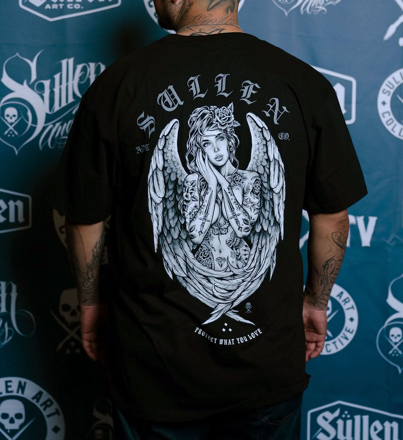 Person wearing a black t-shirt with a white angel design on the back, standing against a branded backdrop.