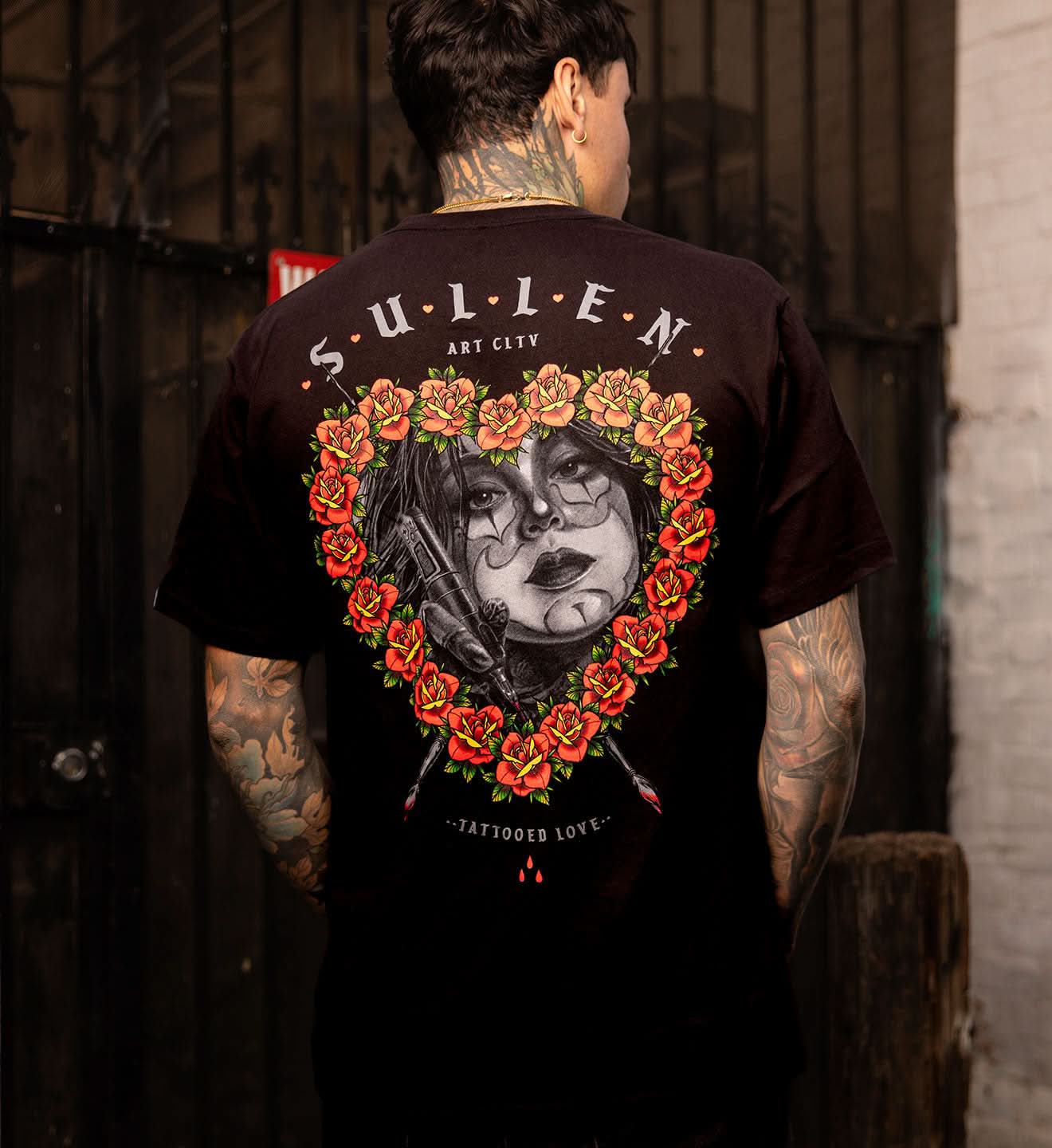 BLACK SULLEN T SHIRT WITH A SOFT GRAPHITE PORTRAIT OF A FEMALE TATTOOER FRAMED WITH TRADITIONAL COLORED ROSES IN THE SHAPE OF A HEART