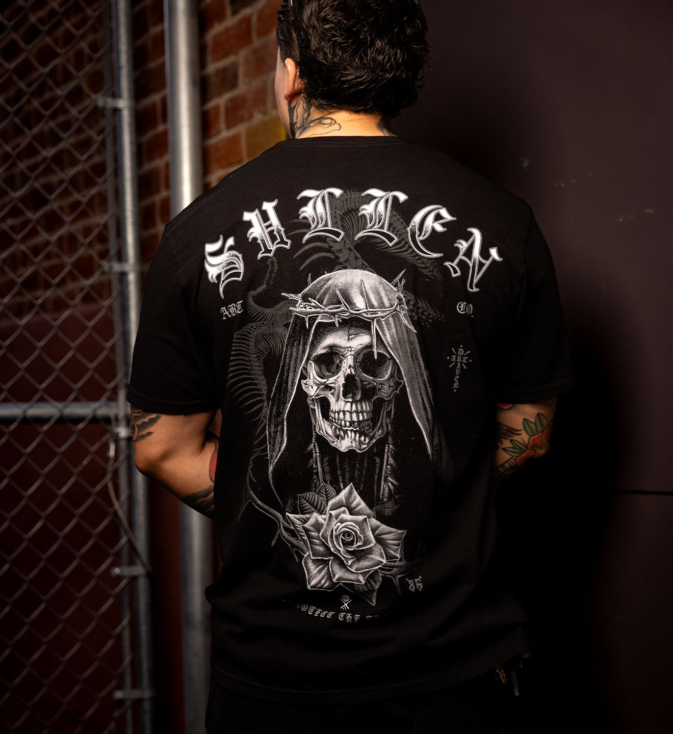 Person wearing a black t-shirt with a skull and rose design on the back, standing against a dark background.